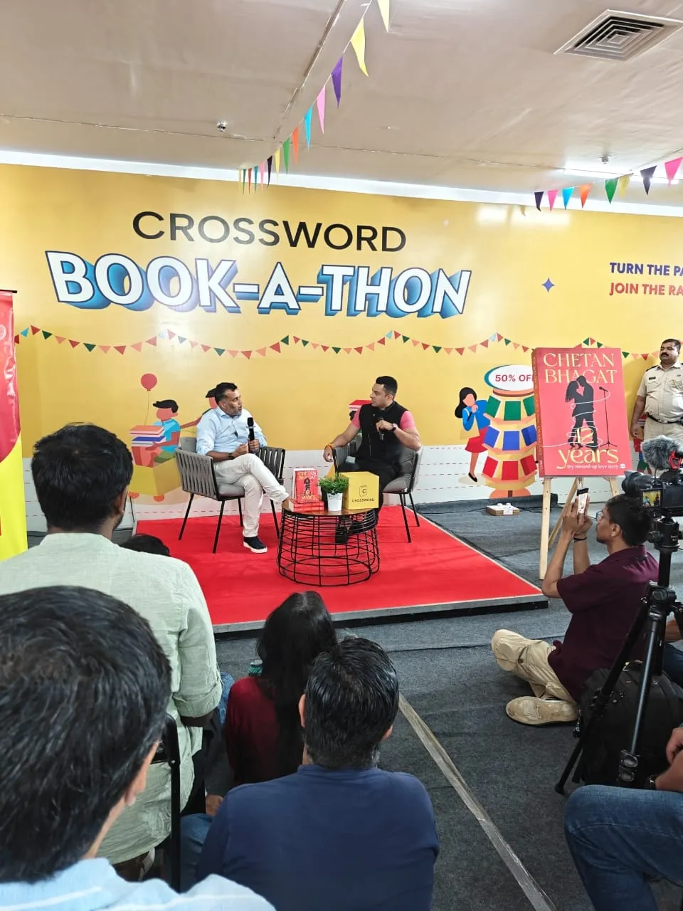 An author session in progress at the Crossword Book-a-thon, with an audience seated and a camera crew filming.