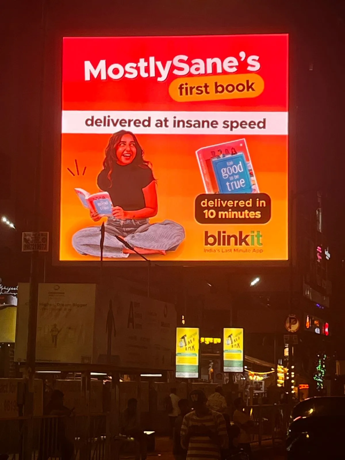 A lit billboard reading “MostlySane’s first book delivered at insane speed — delivered in 10 minutes”, advertising Too Good To Be True on Blinkit.